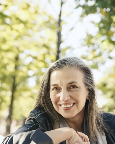 Portrait of a smiling woman with long grey hair outdoors under trees